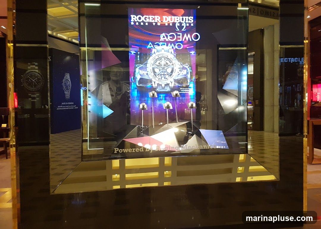 A digital display showcases a Roger Dubuis and Omega collaboration watch. The display features a car and the watches are presented on stands. The text "Powered by Raging Mechanics" is visible at the bottom. The display is set within a mirrored frame, suggesting a high-end retail environment.