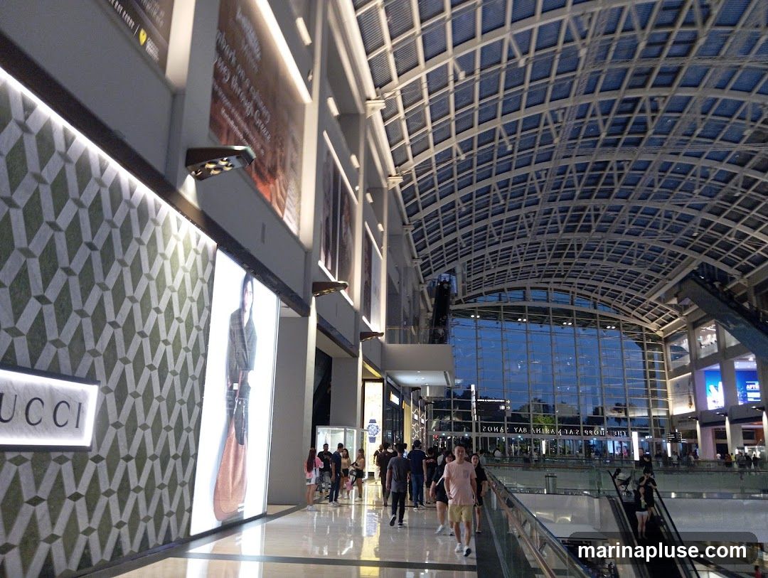 A wide shot of the interior of a large, modern shopping mall. The ceiling is a complex lattice structure, and large windows offer a view of the outside. People are seen walking on the ground floor and on escalators. A prominent display features the word "GUCCI" and a fashion advertisement. The overall impression is one of a spacious and upscale retail environment.