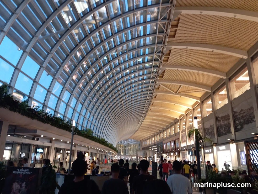 A wide-angle view of a modern indoor shopping mall corridor with a distinctive arched glass ceiling. The ceiling structure is made of metal beams and glass panels, allowing natural light to filter in. On the right side, the corridor features shops with illuminated storefronts and large decorative murals on the walls. People are seen walking along the corridor, suggesting a busy atmosphere. The overall impression is one of a spacious and contemporary retail environment.