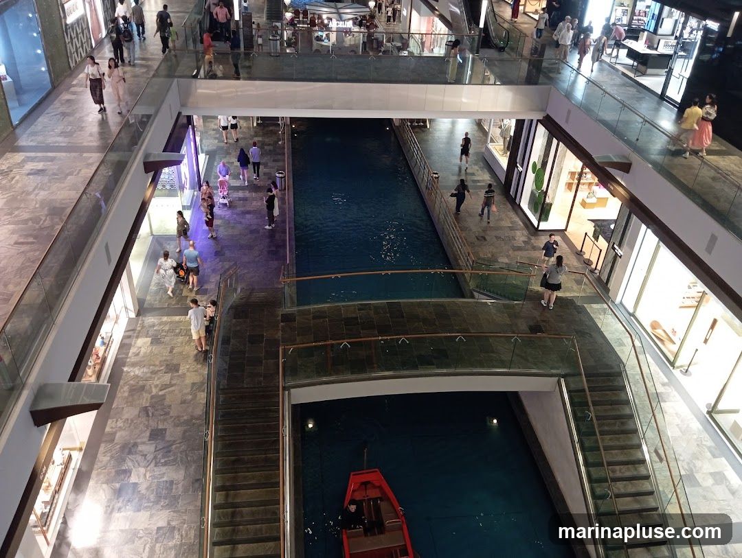 An overhead view of a multi-level indoor shopping mall with a canal running through the center. People are seen walking on the walkways and bridges, and a small red boat is on the canal. Shops with illuminated displays line the canal.