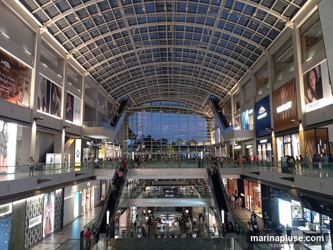 A wide-angle view of the interior of a multi-level shopping mall with a glass ceiling. Escalators and walkways connect different floors, and numerous shops with illuminated displays line the sides. People are seen browsing and walking around. Brands like Dior, Gucci, Chanel, and Hublot are visible on storefronts.