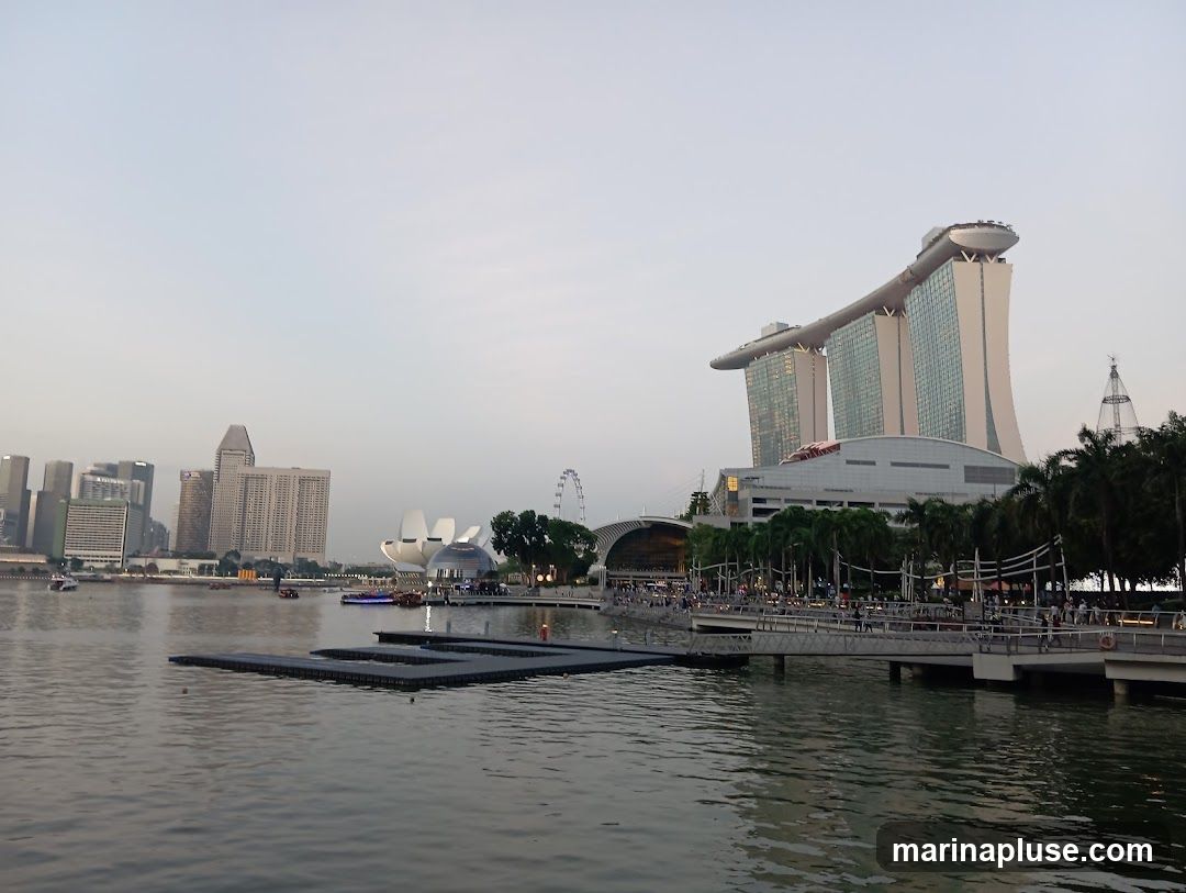 A wide shot of Marina Bay in Singapore, featuring the iconic Marina Bay Sands hotel with its distinctive boat-shaped top. In the background, the city skyline is visible, including various skyscrapers and a Ferris wheel. The foreground shows the calm waters of the bay with a floating platform. The sky is overcast, suggesting it might be early morning or late afternoon. People are seen walking along a promenade in front of the hotel.
