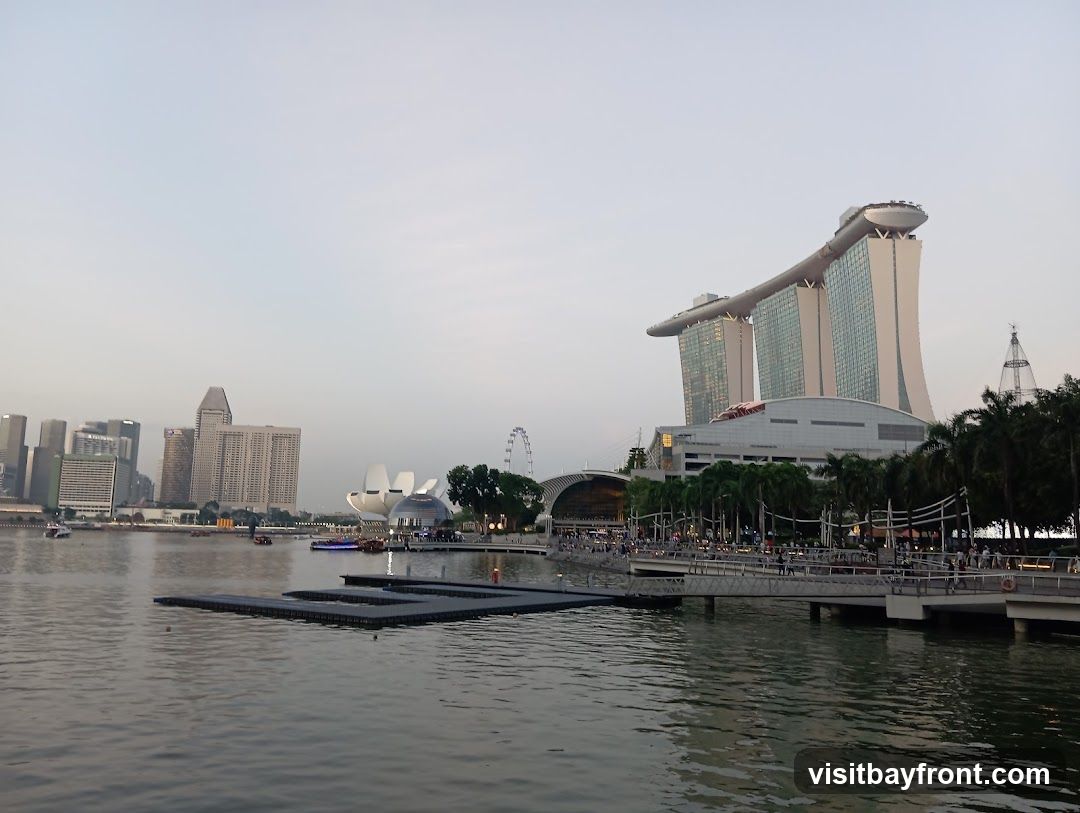 A wide shot of Marina Bay in Singapore, featuring the iconic Marina Bay Sands hotel with its distinctive boat-shaped top. In the background, the city skyline is visible, including various skyscrapers and a Ferris wheel. The foreground shows the calm waters of the bay with a floating platform. The sky is overcast, suggesting it might be early morning or late afternoon. People are seen walking along a promenade in front of the hotel.