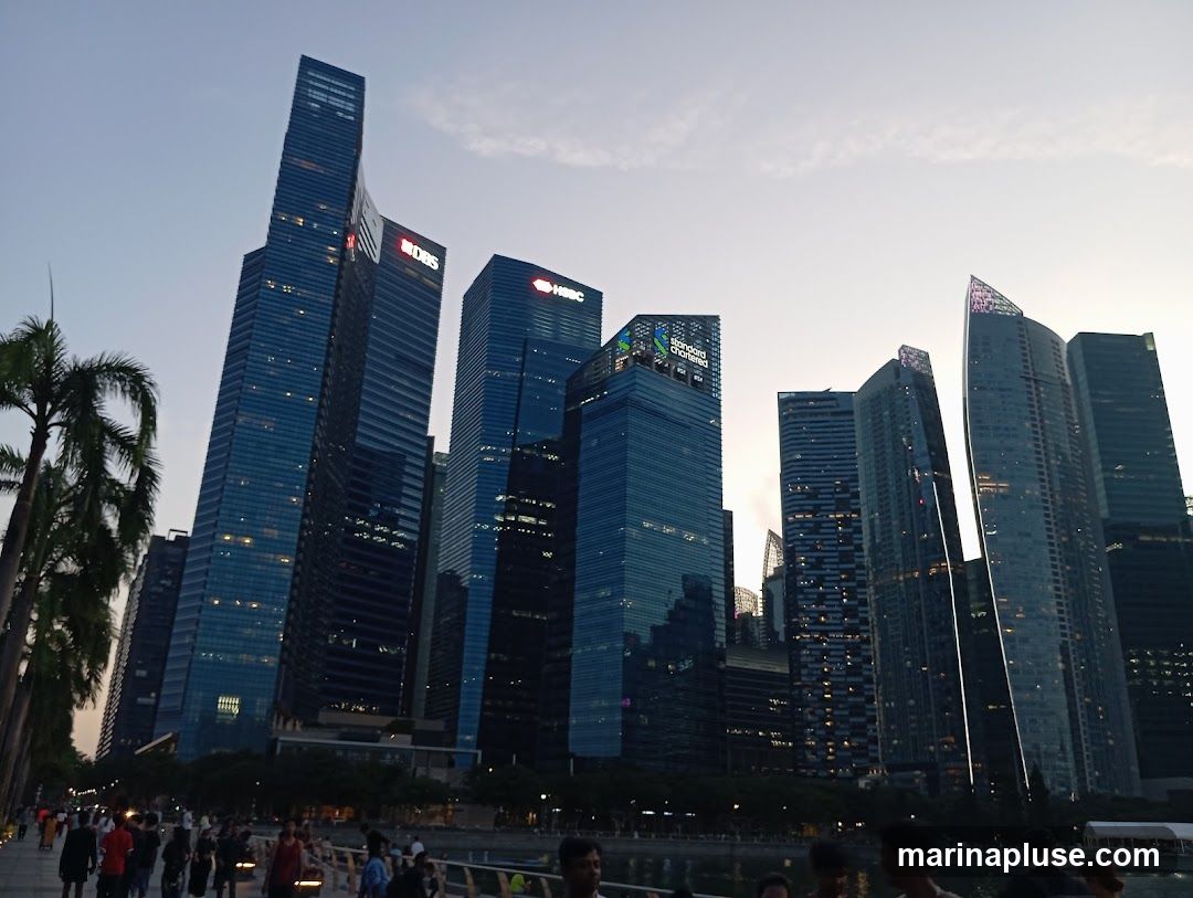 A wide shot of a modern city skyline at dusk, with numerous skyscrapers of varying heights and architectural styles. The buildings are illuminated, with some displaying prominent company logos such as DBS and HSBC. Palm trees line the left side of the frame, and a waterfront promenade with people walking is visible in the foreground. The sky is a soft gradient of blue and white, indicating the transition from day to night.