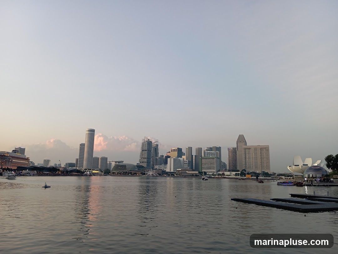 A panoramic view of the Singapore skyline at dusk, with modern skyscrapers and distinctive architecture reflected in the calm waters of a bay. Several boats are visible on the water, and the sky is a soft gradient of pale blue and pink.