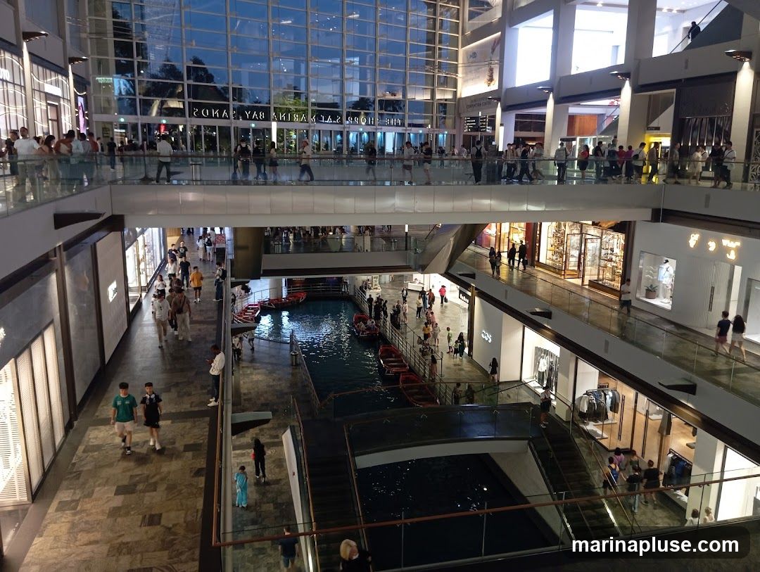 A wide shot of the interior of a large, modern shopping mall. The mall features multiple levels with shops and walkways. In the center, there is a canal with gondola-like boats carrying people. People are seen walking on the upper and lower levels, browsing in shops, and observing the canal. The architecture is contemporary, with large glass windows reflecting the interior lights and the sky outside.