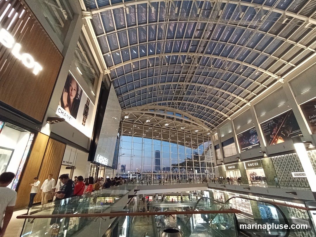 The image shows the interior of a large, modern shopping mall with a distinctive glass and metal arched ceiling. Several high-end stores, including Chanel and Gucci, are visible on the upper levels, with their prominent signage. Advertisements for events like the "Singapore Night Race 2024" are displayed on the walls. People are seen browsing and walking around the mall, with escalators and railings indicating multiple levels. The overall impression is one of a bustling, upscale retail environment with a view of the city skyline through the large glass facade at the far end.