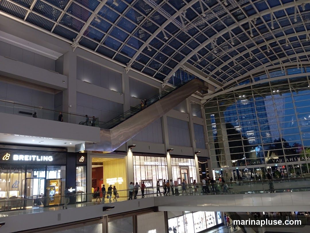 The image shows the interior of a large, modern shopping mall with a distinctive glass and metal roof structure. Escalators are visible, connecting different levels of the mall. Several storefronts are visible, including one with the prominent "BREITLING" logo. People are seen walking on the elevated walkways and escalators. The exterior of a tall building is reflected in the large glass windows of the mall.