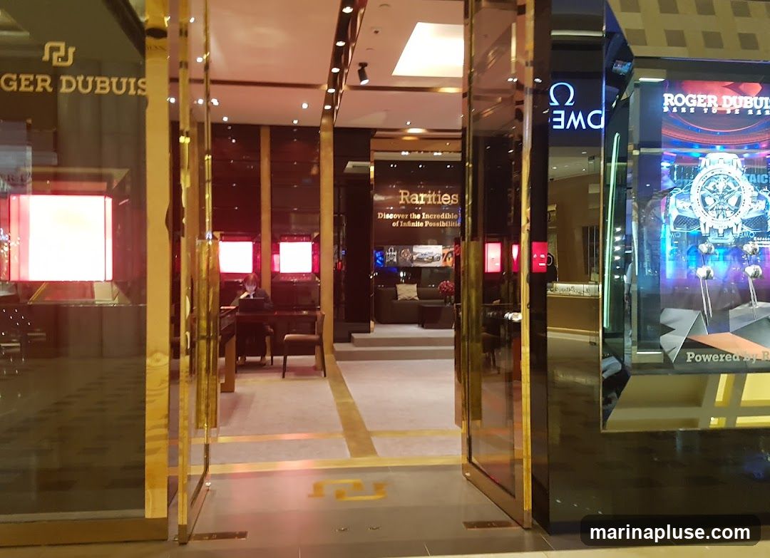 The entrance to a luxury watch store with a gold-trimmed facade. The store features large display windows showcasing watches and branded signage. Inside, a person is seated at a desk, and a lounge area is visible.