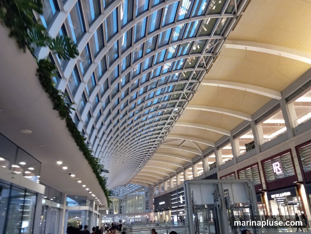 An interior view of a spacious, modern shopping mall featuring a distinctive arched skylight made of glass panels and a white grid structure. Lush green plants are draped along the upper edge of the walkway. The mall's interior is illuminated by natural light filtering through the skylight and by artificial lights. On the right side, storefronts with illuminated signs are visible, including one with a prominent 'R' logo. People are seen walking through the mall, indicating a busy commercial environment. The architecture emphasizes clean lines and a bright, airy atmosphere.