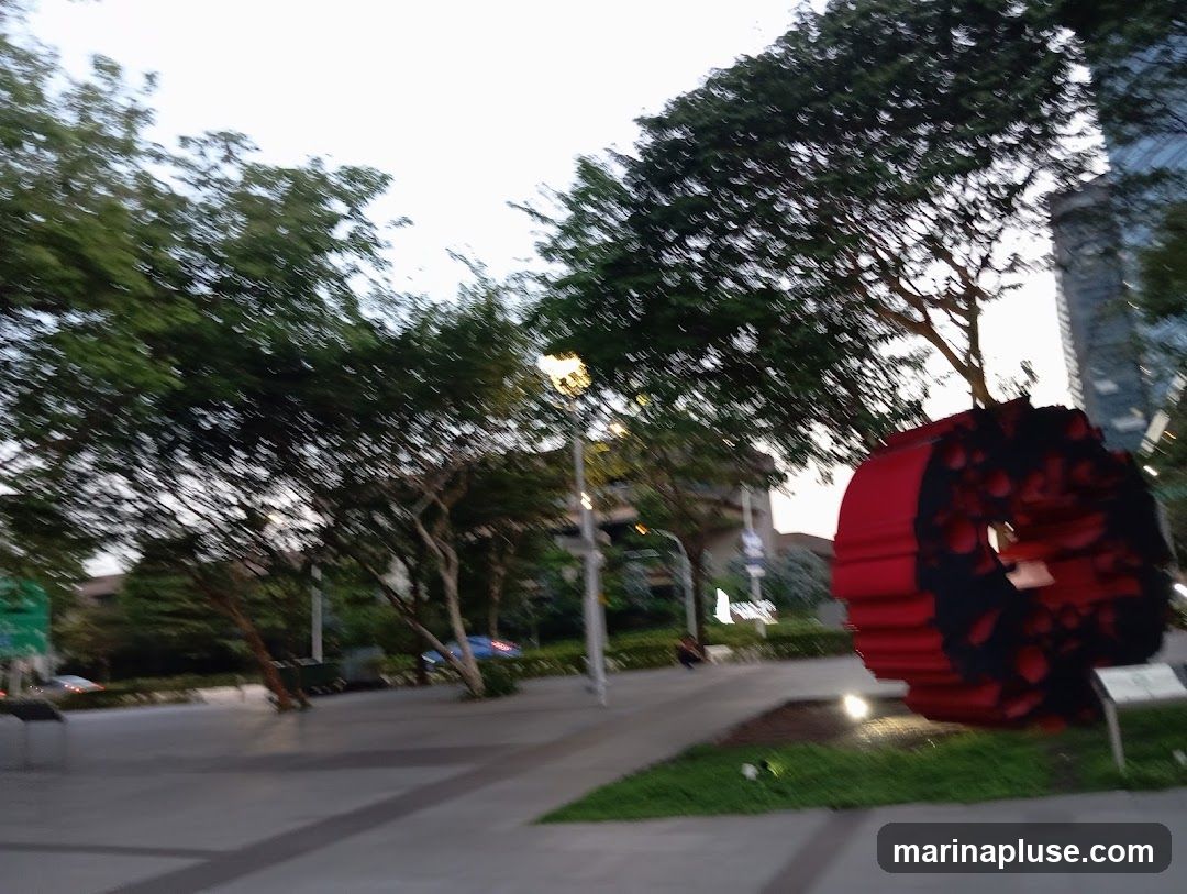 A large, circular, red and black sculpture with petal-like designs is displayed outdoors on a paved area with grass. Trees and buildings are visible in the background, and the sky is overcast. The image is blurry, suggesting motion or low light conditions.