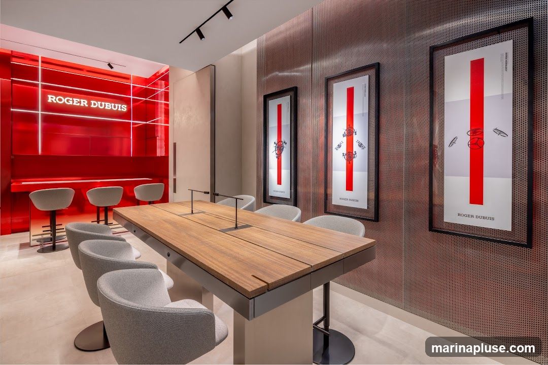 The interior of a modern luxury watch store, featuring a prominent red wall with the "ROGER DUBUIS" logo illuminated in white. A long wooden table with chairs is set up in the center, with bar stools along the red counter. The walls are decorated with framed posters showcasing watch components and the brand name.