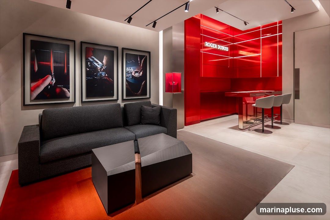 A sleek and modern interior of a luxury watch boutique. A dark grey sectional sofa is positioned on an orange rug, with two geometric coffee tables in front of it. Three framed artworks depicting watchmaking processes hang on the wall behind the sofa. To the right, a striking red wall features the "ROGER DUBUIS" logo illuminated by white LED strips. A red display case sits on a metallic pedestal. A high, reflective table with three light grey bar stools is set against the red wall. The floor is tiled in a light, neutral tone.