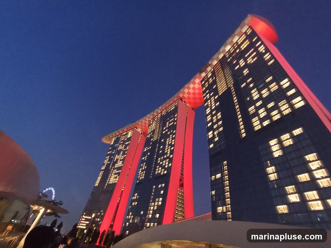 A low-angle shot of the Marina Bay Sands hotel in Singapore at dusk. The iconic building, with its three towers connected by a sky-high platform, is illuminated by red lights and the warm glow of its many windows. The deep blue of the twilight sky provides a dramatic backdrop. In the distance, a Ferris wheel can be seen.