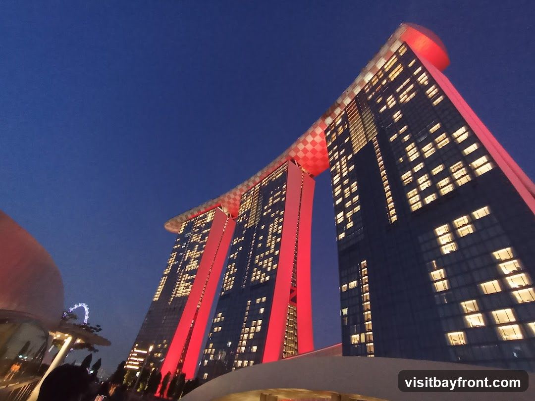 A low-angle shot of the Marina Bay Sands hotel in Singapore at dusk. The iconic building, with its three towers connected by a sky-high platform, is illuminated by red lights and the warm glow of its many windows. The deep blue of the twilight sky provides a dramatic backdrop. In the distance, a Ferris wheel can be seen.