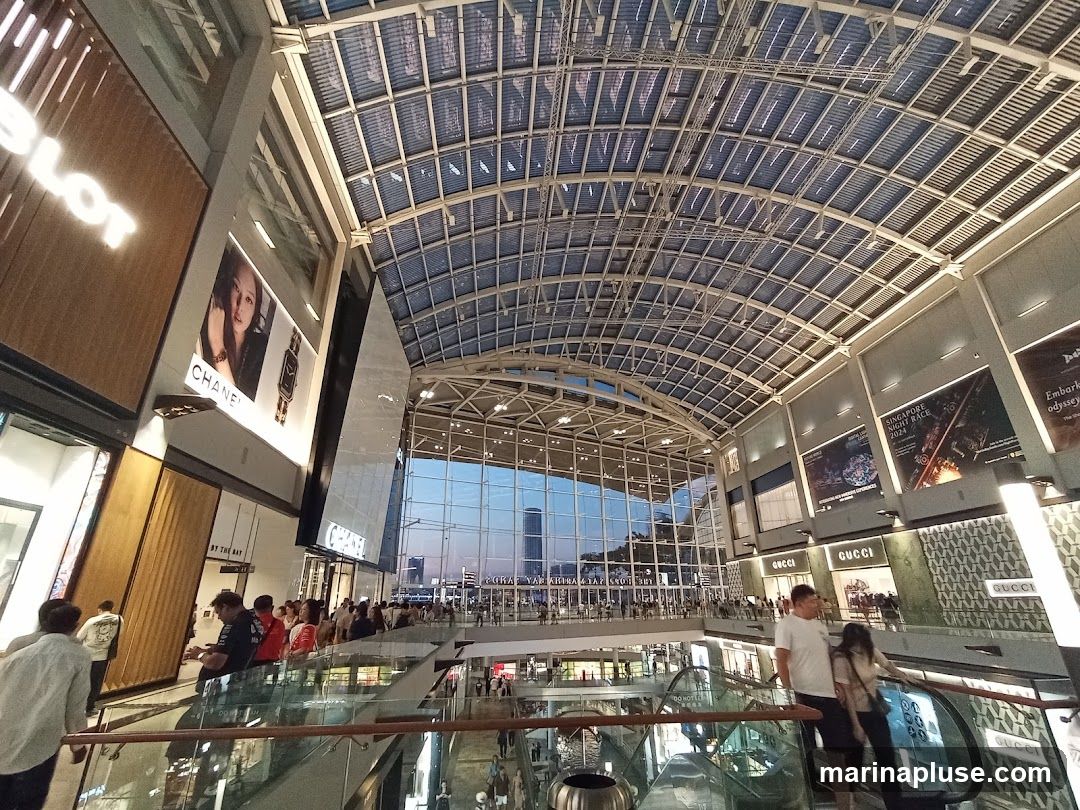 The image shows the interior of a large, modern shopping mall with a high, arched glass ceiling. Several shops are visible on multiple levels, including HUBLOT, CHANEL, and GUCCI. Advertisements and posters are displayed on the walls. People are seen walking around and using escalators. The overall atmosphere is bright and spacious, with a view of the city skyline through the glass facade in the distance.