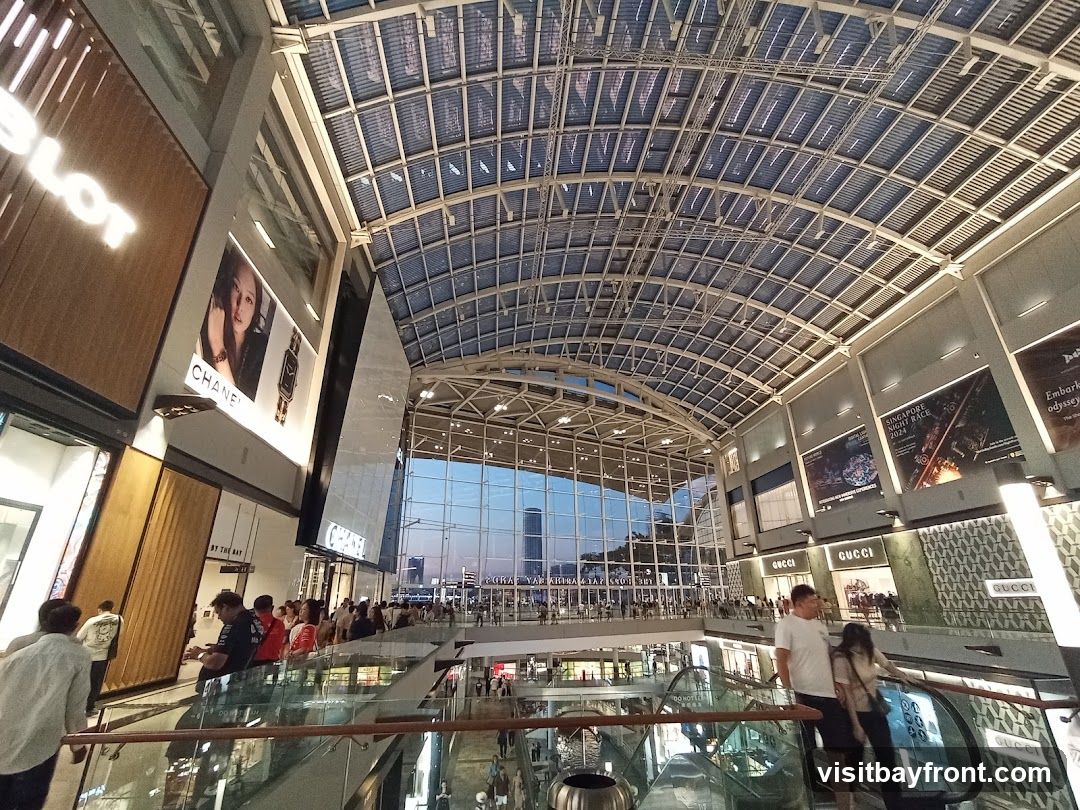 The image shows the interior of a large, modern shopping mall with a high, arched glass ceiling. Several shops are visible on multiple levels, including HUBLOT, CHANEL, and GUCCI. Advertisements and posters are displayed on the walls. People are seen walking around and using escalators. The overall atmosphere is bright and spacious, with a view of the city skyline through the glass facade in the distance.
