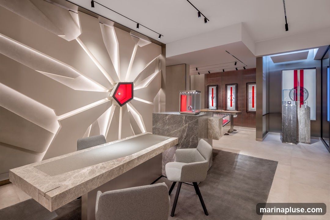 The interior of a modern, upscale watch store. The walls feature a striking geometric design with integrated lighting, and a prominent red accent piece. A large marble-topped table with chairs is in the foreground. Display cases with watches are visible throughout the store, along with framed artwork on the back wall. The overall ambiance is sophisticated and minimalist.