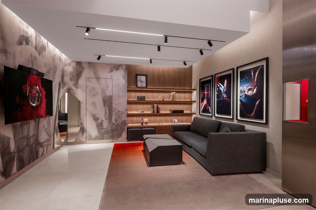 A contemporary showroom features a large television displaying an artistic image, a grey sectional sofa, a modern coffee table, and a bookshelf filled with books and decorative items. Three framed artworks depicting watchmaking are displayed on the wall. The space has a sleek, minimalist design with track lighting and a gradient rug.