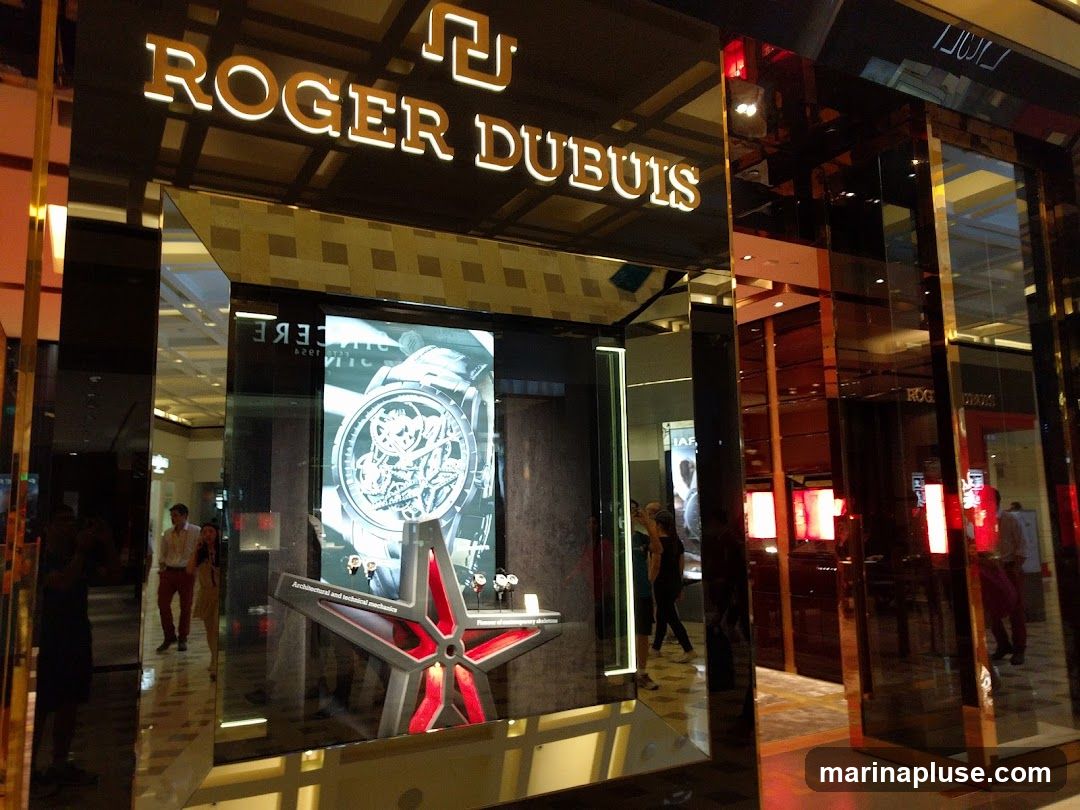 The exterior of a Roger Dubuis watch store. The prominent signage "ROGER DUBUIS" is illuminated in gold lettering above the entrance. A large digital display inside showcases a detailed image of a luxury watch, with a star-shaped display stand in front of it. The store features a modern, sleek design with reflective surfaces and glass panels. People can be seen inside and reflected in the glass.