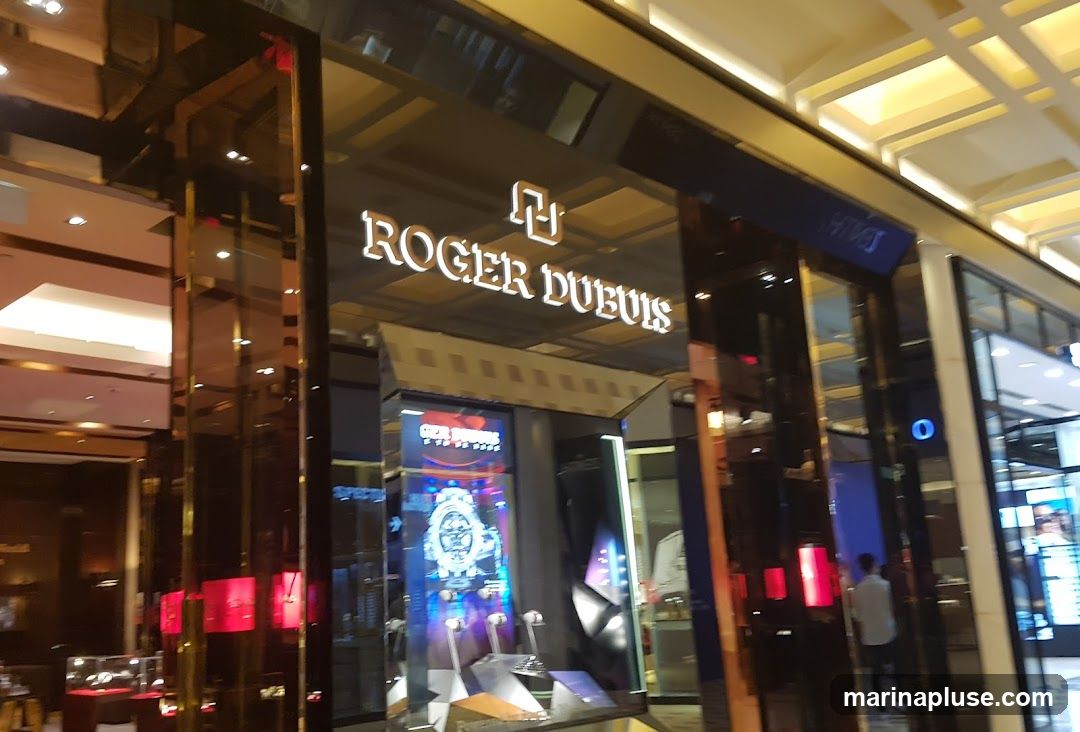 The exterior of a Roger Dubuis watch store. The store has a large, illuminated sign with the brand name "ROGER DUBUIS" and a logo above the entrance. The storefront features large glass windows displaying watches and promotional materials. The interior is visible through the windows, showing display cases and lighting.