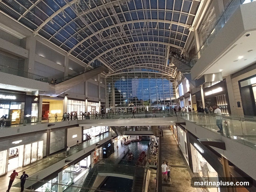 A wide-angle view of the interior of a large, multi-level shopping mall. The architecture features a prominent glass and metal roof structure, allowing natural light to filter in. Escalators and walkways connect different levels, and numerous shops with illuminated storefronts line the corridors. In the lower section, a canal with gondolas is visible, adding a unique element to the mall's design. People are seen browsing shops, walking on the elevated walkways, and riding escalators. The overall atmosphere is one of a bustling commercial and leisure space.