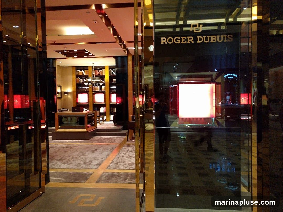 The image shows the exterior of a luxury watch store. The prominent signage above the entrance reads "ROGER DUBUIS". The storefront features large glass windows and doors, reflecting the interior and the street. Inside, the store is elegantly designed with display cases, bookshelves, and soft lighting, showcasing a sophisticated ambiance.