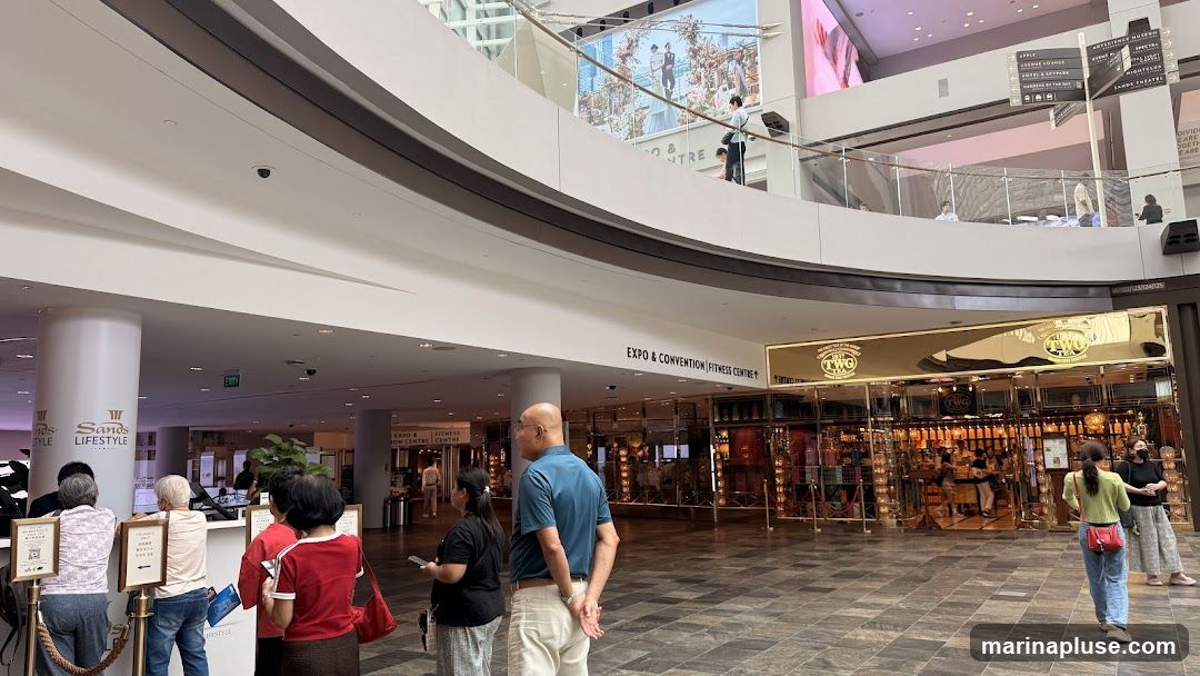 The image shows the interior of a modern shopping mall. In the foreground, people are queuing at a counter with signs that say "Sands Lifestyle". In the background, there is a prominent shop with a golden facade and the TWG logo, which appears to be a tea shop. The shop is filled with shelves displaying numerous tea containers. Further in the background, there are signs indicating directions to "Expo & Convention Fitness Centre" and other locations like "Apple", "ArtScience Museum", and "Jardine Theatre". A large digital screen displays an advertisement.