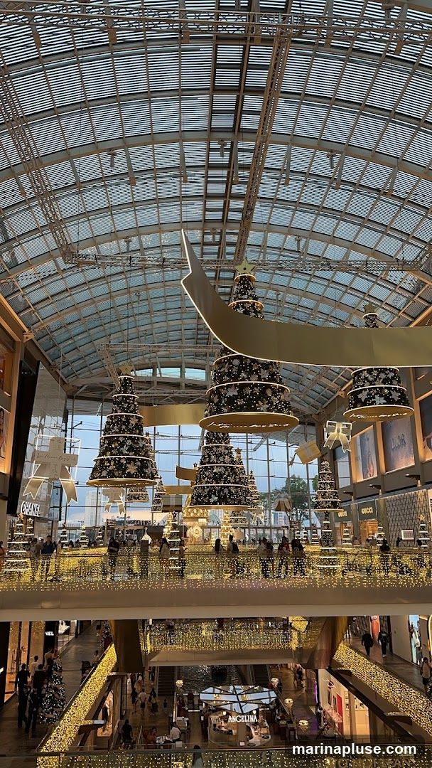 A wide shot of the interior of a shopping mall decorated for Christmas. Large, tiered Christmas trees are suspended from the ceiling, adorned with lights and ornaments. The mall features multiple levels with escalators and walkways, all decorated with strings of warm-toned lights. Various high-end stores, including Chanel, Gucci, and Dior, are visible on the lower levels. People are seen browsing and walking through the mall. The ceiling is a glass and metal structure, allowing natural light to filter in.