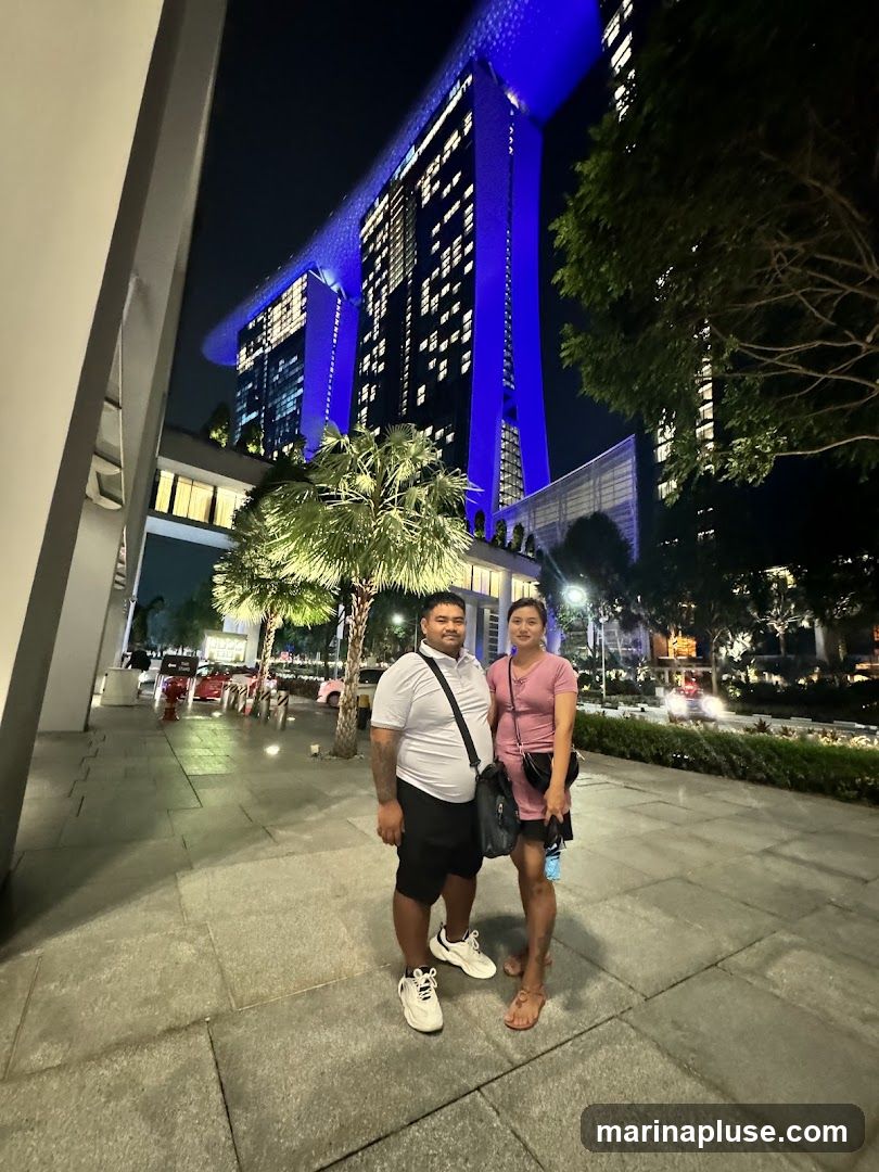 A couple stands on a paved walkway at night, with the iconic Marina Bay Sands hotel illuminated in blue in the background. The man is wearing a grey polo shirt and black shorts, while the woman is in a pink dress. Palm trees and other greenery are visible around them. The building's distinctive architecture, with its three towers connected by a sky park, is prominently displayed.