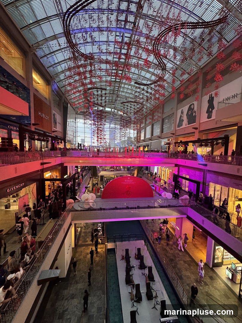An interior view of a multi-level shopping mall, illuminated by pink and purple lights. Red, intricate decorations hang from the glass ceiling, creating a festive atmosphere. Various high-end stores like HUBLOT, CHANEL, DIOR, and GIVENCHY are visible on different levels. People are seen browsing and walking around the mall. In the center, there is a water feature with chairs and musical instruments set up, possibly for a performance. A large red circular display with Chinese characters is also prominent.