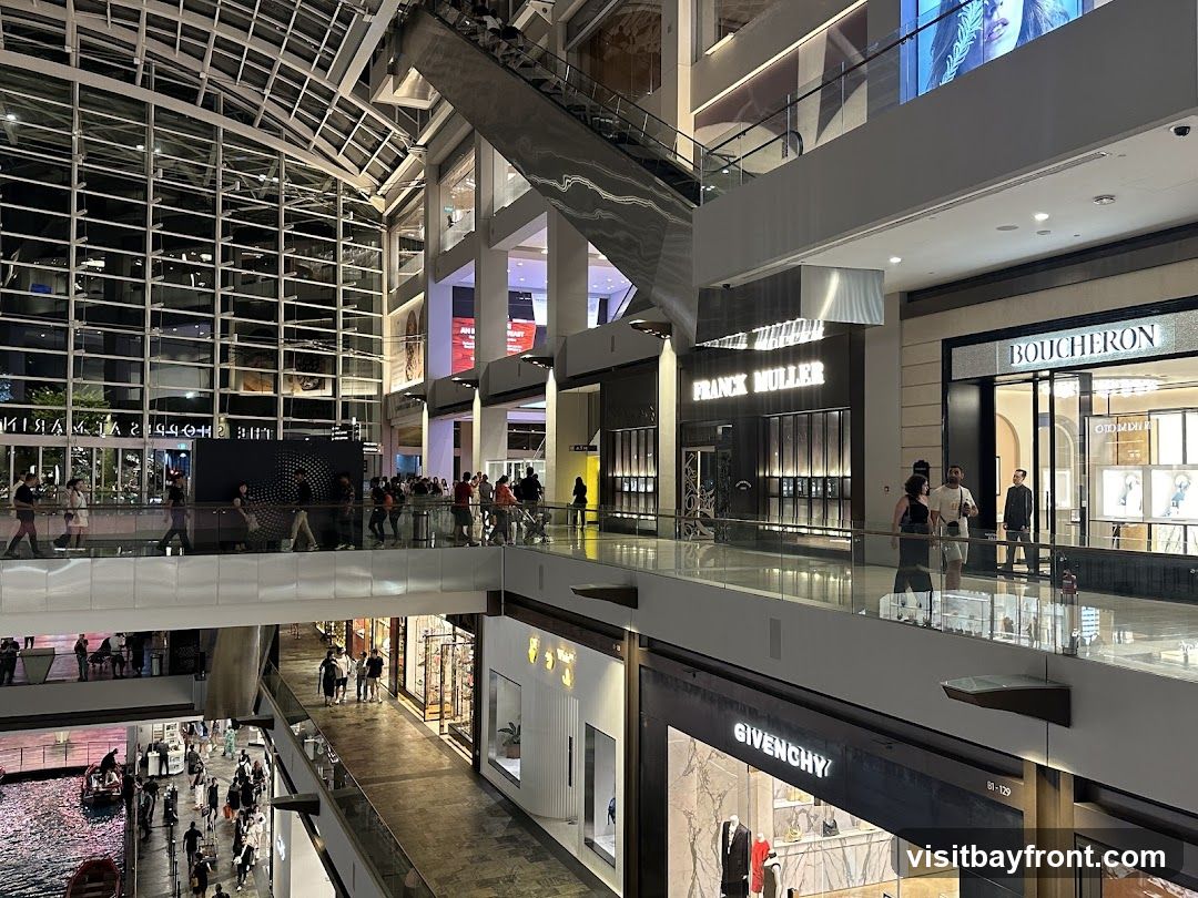 The image shows the interior of a large, multi-level shopping mall. Escalators and walkways connect different floors. Several high-end retail stores are visible, including Franck Muller, Boucheron, and Givenchy, with their prominent signage. People are seen walking around the mall and browsing in the stores. The architecture features a glass ceiling and modern design elements. Below, a canal with boats can be seen, suggesting a unique waterfront location within the mall.