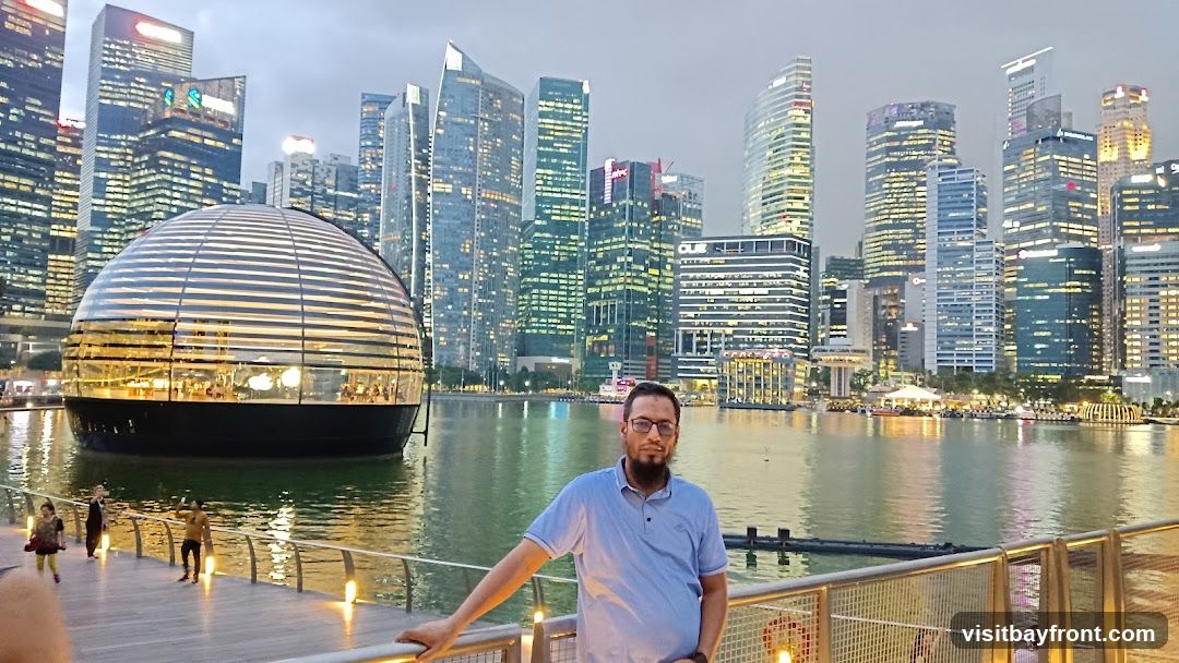 A man with a beard and glasses stands on a wooden walkway, leaning on a railing. Behind him, a distinctive spherical glass building with horizontal accents is situated on the water. The background features a dense skyline of modern skyscrapers, illuminated by artificial lights, reflecting on the water. Several people are visible on the walkway and in the distance. The overall scene suggests an urban waterfront setting at dusk.
