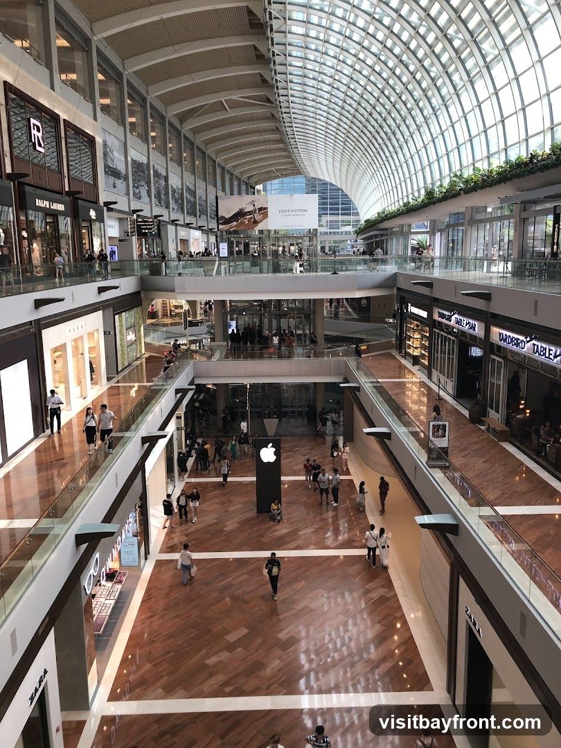 An aerial view of the interior of a large, modern shopping mall. The mall features a distinctive curved glass roof, multiple levels with escalators and walkways, and polished wooden floors. Various retail stores are visible on different levels, including Ralph Lauren, Gucci, Zara, and Louis Vuitton. People are seen browsing and walking through the mall. An Apple Store is prominently displayed with its logo.