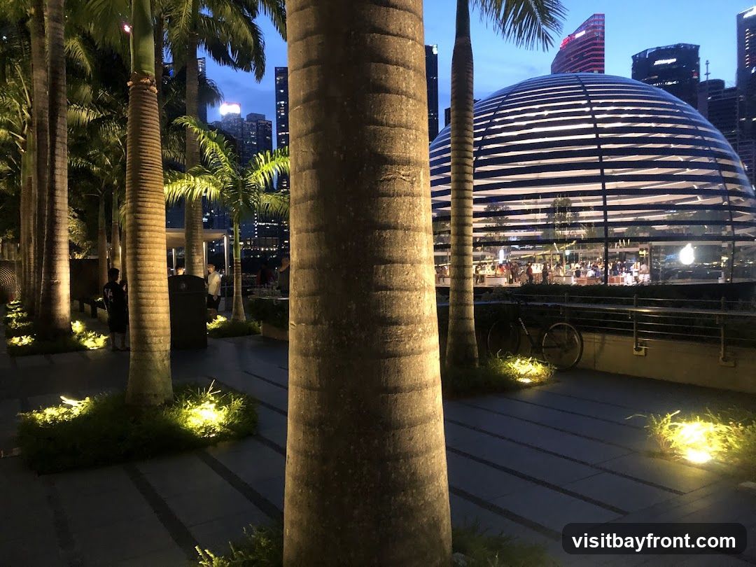A striking, modern glass dome building with horizontal white accents is illuminated at dusk, set against a backdrop of city skyscrapers. Palm trees line a walkway in the foreground, with soft lighting illuminating the grass and paving stones. People are visible inside the dome, suggesting it is a public space or store. A bicycle is parked near the building.