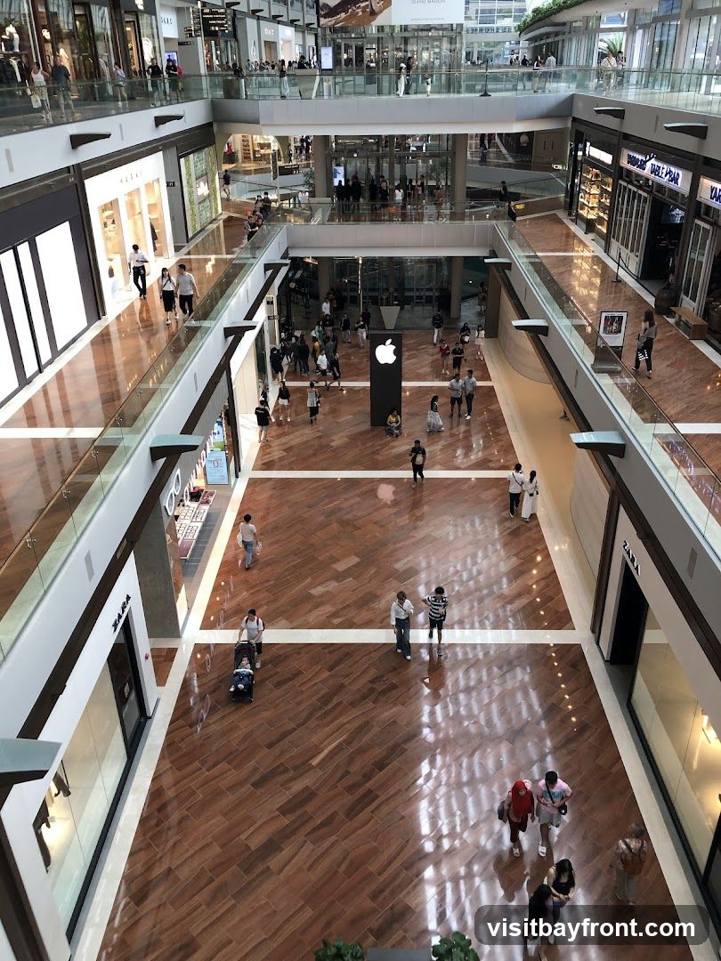 An overhead view of a busy shopping mall with multiple levels. Shoppers are seen walking on the polished wooden floors and along the balconies. Various storefronts are visible, including Zara and what appears to be an Apple store with its iconic logo displayed prominently. The mall features modern architecture with glass railings and bright lighting.