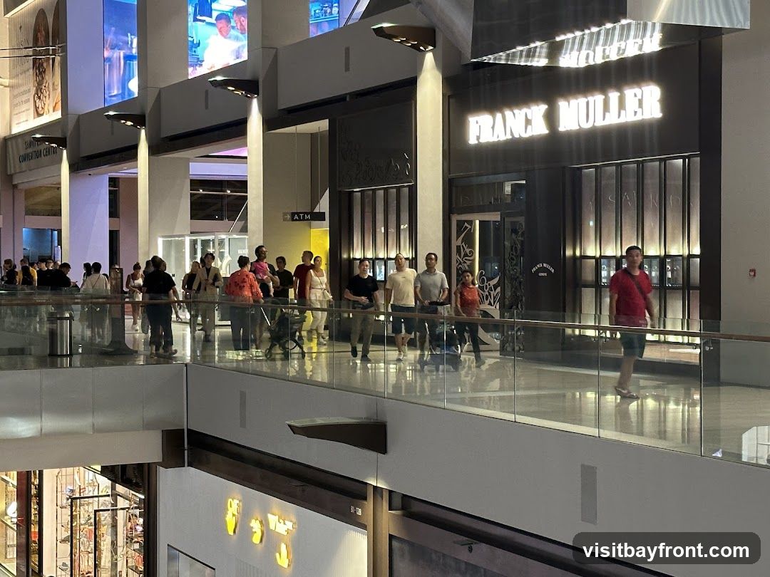 A well-lit storefront for Franck Muller, a luxury watch brand, is visible in a modern indoor shopping mall. The store features a dark facade with illuminated signage displaying the brand name. Several people are walking on a walkway in front of the store, with a glass railing separating them from the lower level. Other shops and displays are visible in the background, along with large screens showing advertisements.