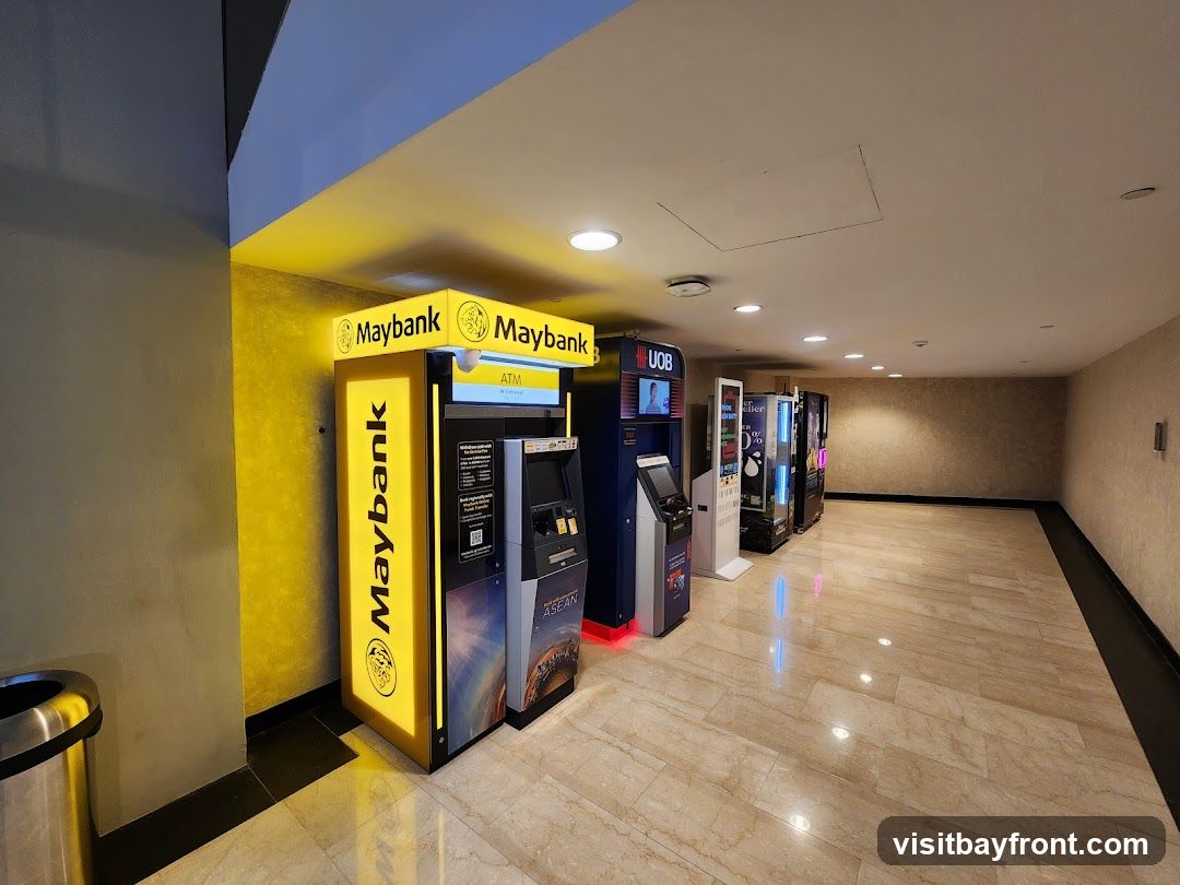 A hallway with several ATMs and vending machines. The first ATM is branded with 'Maybank' in large yellow lettering. Next to it is a UOB ATM. Further down the hall are several colorful vending machines. The floor is tiled with a marble pattern, and the walls are a neutral beige color.