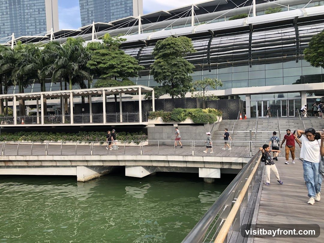 A wide shot of a modern building with a glass facade, surrounded by lush greenery and palm trees. People are seen walking along a wooden promenade next to a body of green water. The building has multiple levels and a distinctive architectural style with white supporting structures.