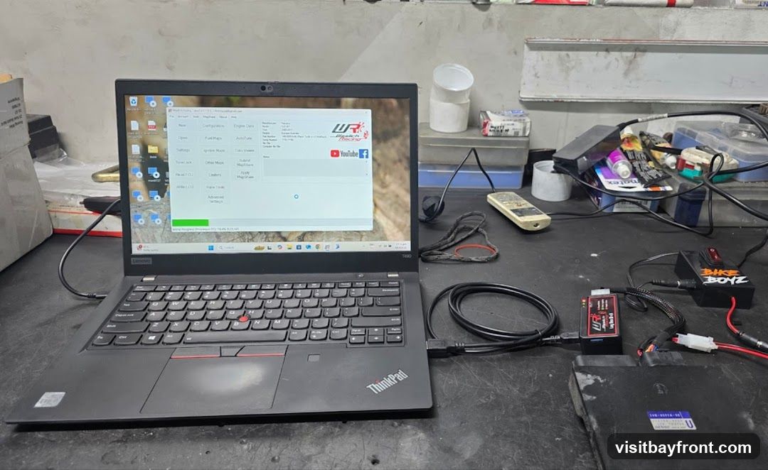 A laptop computer is connected to electronic devices, likely for tuning a motorcycle's Engine Control Unit (ECU). The laptop screen displays software with options for fuel maps, ignition maps, and other tuning parameters. A device labeled 'BIKE BOYZ' is also visible, along with various cables and connectors. The setup appears to be on a workbench in a workshop environment.