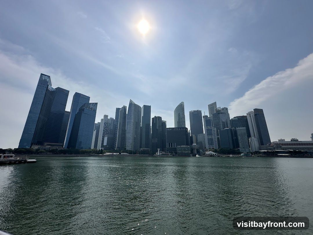 A wide shot of a modern city skyline with numerous skyscrapers of varying heights and architectural styles, viewed across a body of water. The sun is high in the sky, casting a bright glare and creating reflections on the water's surface. The sky is mostly clear with some wispy clouds. The buildings are densely packed, suggesting a major urban center.