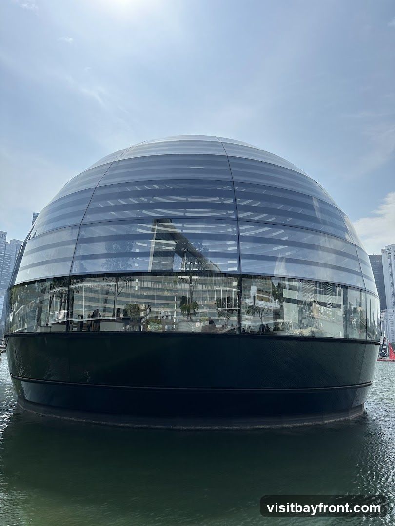 A modern, spherical building with a glass facade sits on the water. The building has a dark, opaque base and a transparent upper section that reveals the interior, including trees and seating areas. The reflection of the sky and surrounding buildings can be seen on the glass.