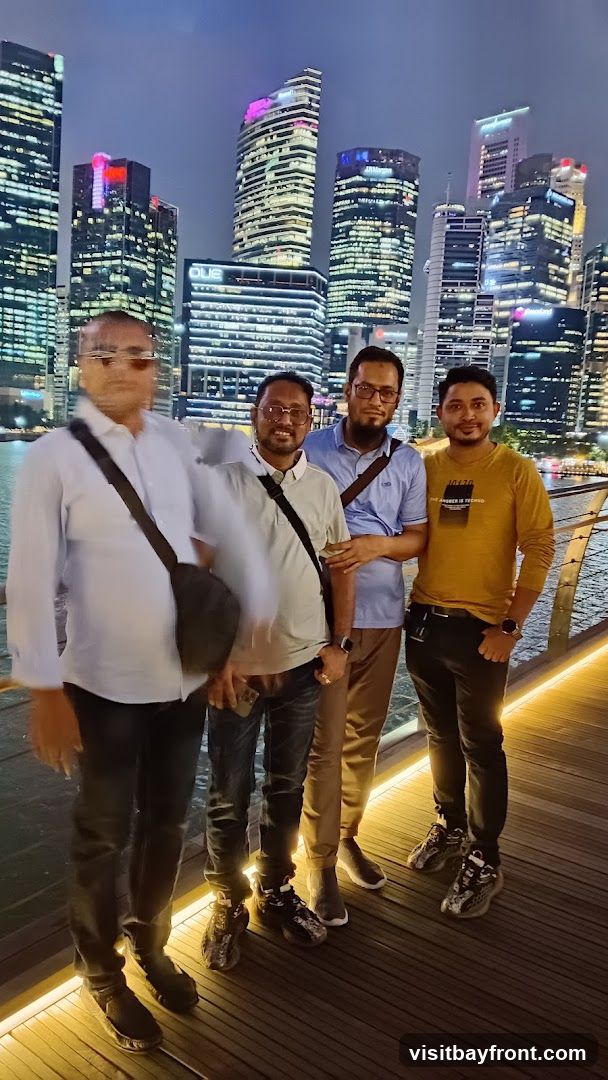 Four men are standing on a wooden walkway at night, with a city skyline in the background. The buildings are illuminated with many lights, and some have colored lights. The men are casually dressed. The man on the far left is wearing a light-colored collared shirt and dark pants. The second man from the left is wearing a light-colored t-shirt, dark jeans, and a crossbody bag. The third man from the left is wearing a blue collared shirt, tan pants, and a crossbody bag. The man on the far right is wearing a yellow long-sleeved shirt with text and dark pants. They are all looking towards the camera.
