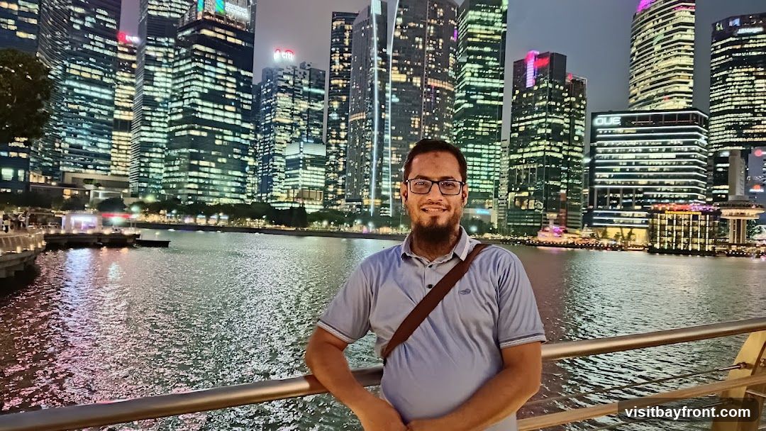A man with glasses and a beard smiles at the camera, wearing a light blue polo shirt and a brown crossbody bag. He is standing on a walkway with a railing in the foreground. Behind him, a body of water reflects the lights of a modern city skyline at night. The skyscrapers are illuminated with numerous windows, creating a vibrant and dazzling backdrop.