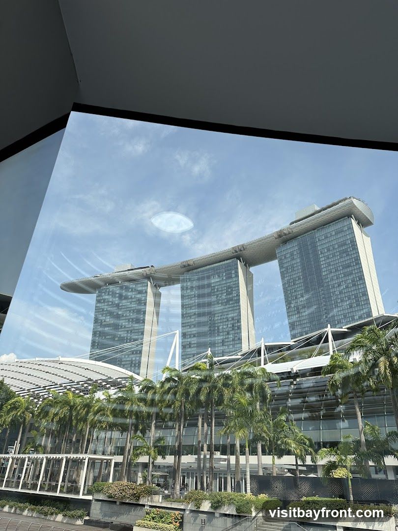 A view of the Marina Bay Sands hotel in Singapore, with its iconic three towers connected by a sky-high platform. Palm trees line the foreground, and the sky is partly cloudy. The image appears to be taken from inside a building, looking out through a window.