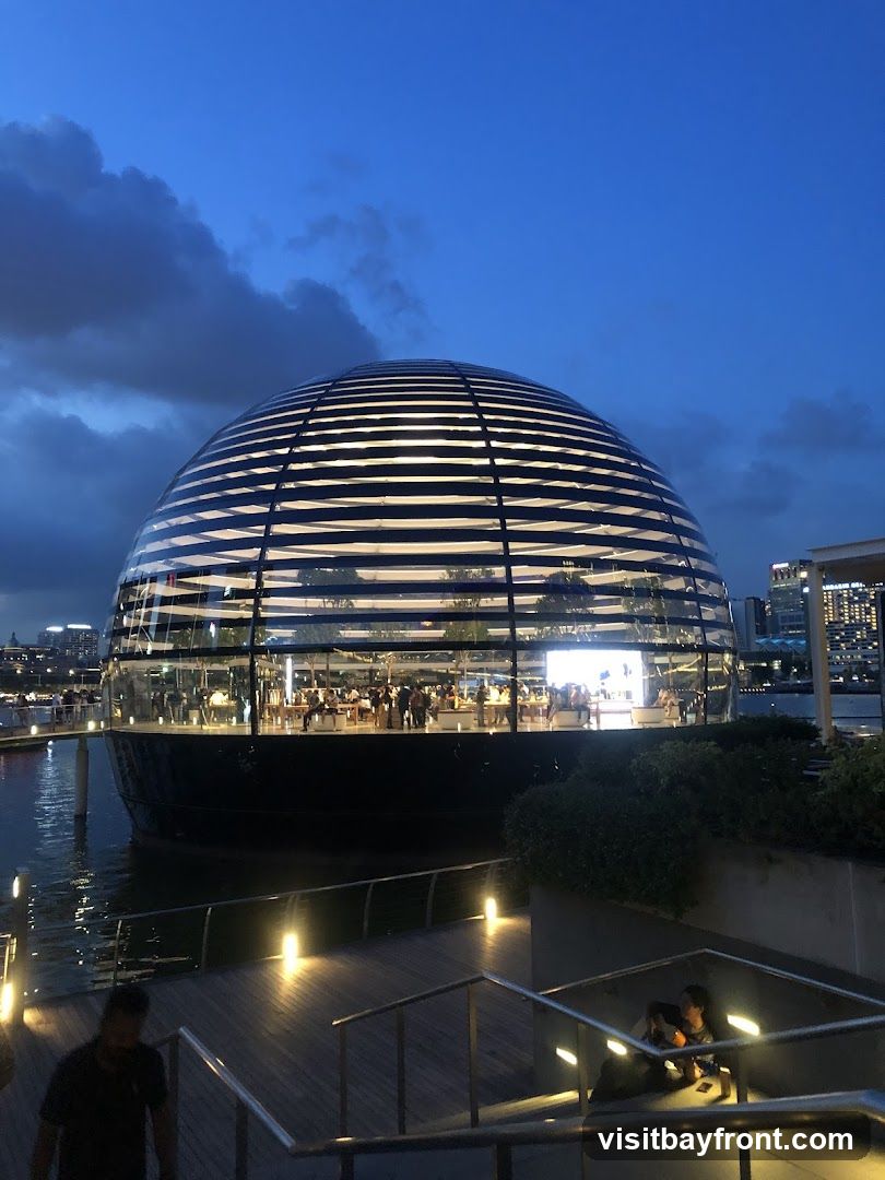 A spherical glass Apple Store is illuminated at dusk, situated on the water. People are visible inside the store, browsing and interacting. A walkway with railings and lighting leads towards the entrance.