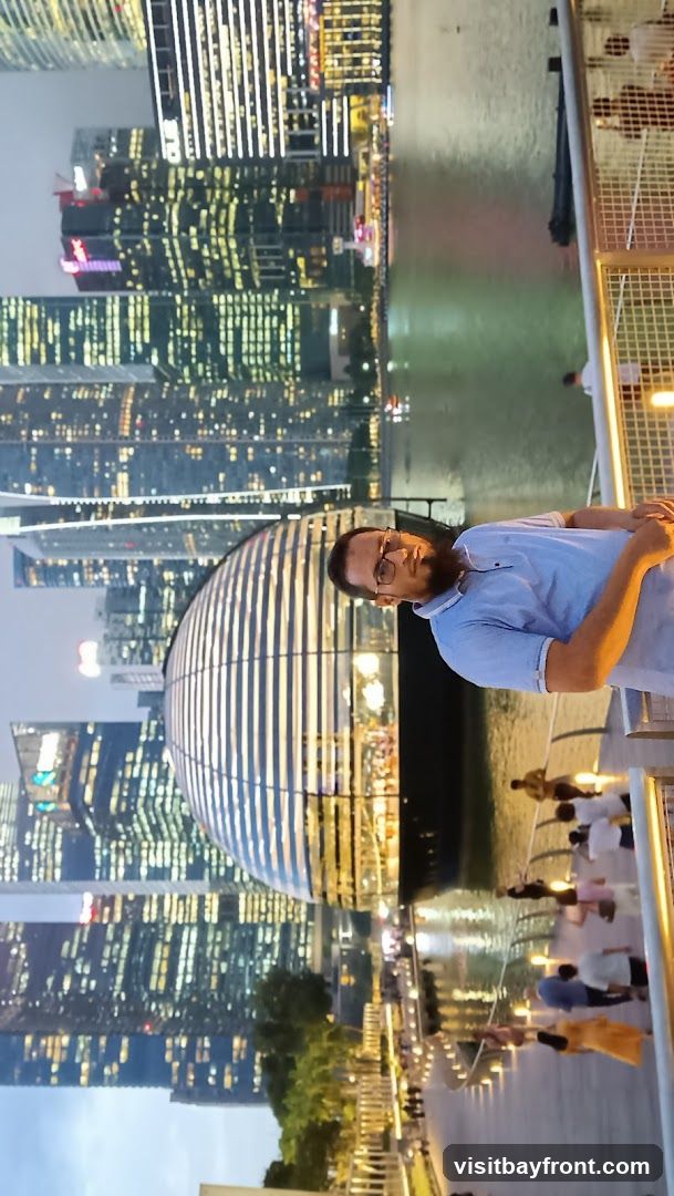 A man wearing glasses and a light blue polo shirt stands on a walkway, posing for a photo. Behind him, a modern city skyline is illuminated at dusk, with numerous skyscrapers featuring bright windows. A distinctive domed building with horizontal stripes is prominent in the foreground. The walkway is lined with railings and lights, and several other people are visible in the background, walking along the waterfront.