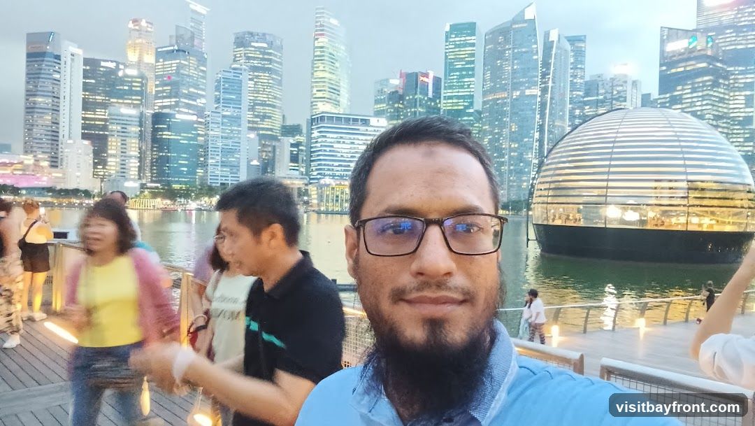 A man takes a selfie with a blurred background of people walking on a boardwalk. Behind them, a modern, spherical glass building sits on the water, reflecting the city skyline. The city skyline is illuminated with many lights, suggesting it is dusk or evening.