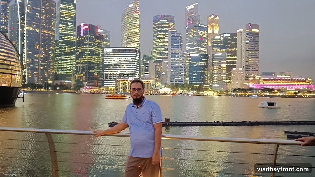 A man stands on a railing overlooking a body of water with a cityscape of illuminated skyscrapers in the background. The sky is overcast, suggesting it is dusk or evening. Several boats are visible on the water, and some buildings in the cityscape are lit with colorful lights.