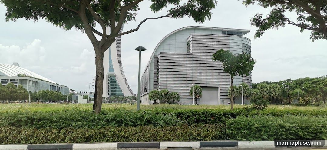 A wide shot of several modern buildings with distinctive architectural designs, including a curved facade and a tall, slender tower. The buildings are surrounded by lush green landscaping, with trees and bushes in the foreground and middle ground. The sky is overcast.